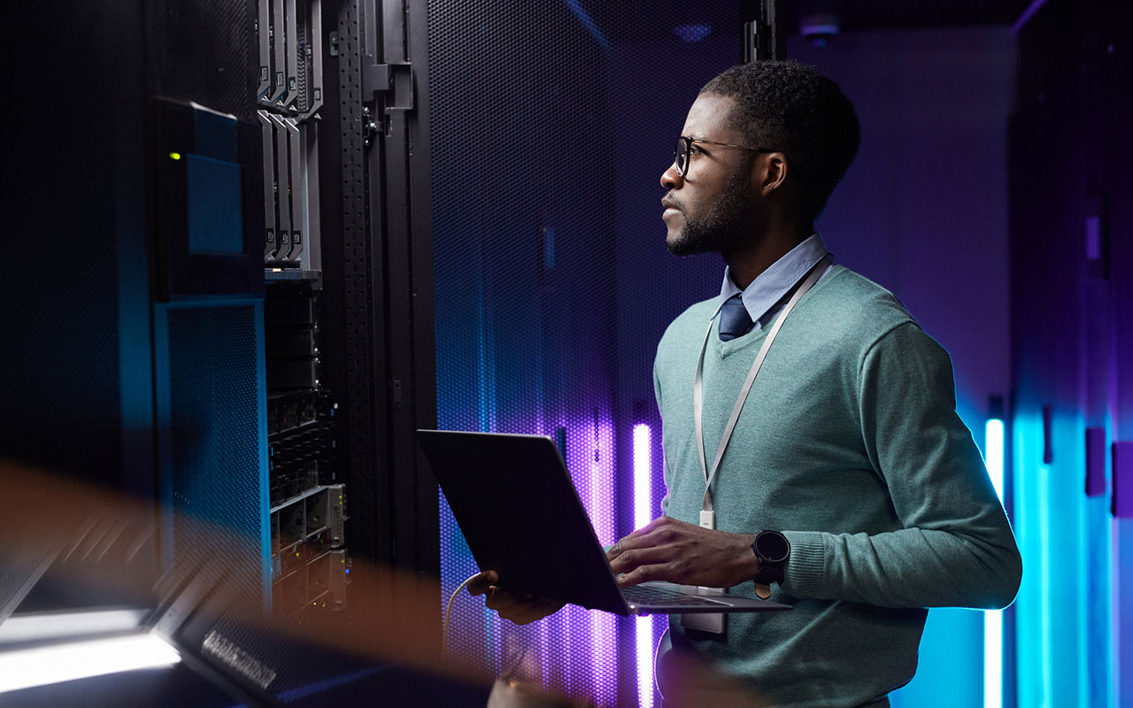 African American data engineer holding laptop while working with supercomputer in server room
