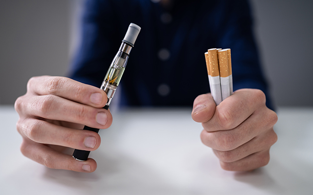 A man holds an e-cigarette in one hand and traditional cigarettes in the other.